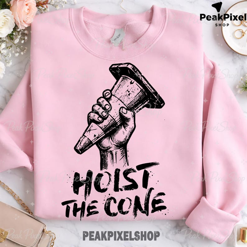 Pu&ograve; includere: Felpa rosa chiaro con una grafica nera di una mano che tiene un cono stradale. Le parole "HOIST THE CONE" sono stampate sotto in un carattere audace e invecchiato. Il nome del negozio, PEAKPIXELSHOP, &egrave; stampato in basso.