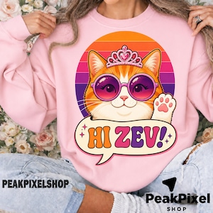 Könnte beinhalten: Rosa Sweatshirt mit einer Cartoon-Katze, die eine Sonnenbrille und ein Diadem trägt, mit einer Sprechblase mit der Aufschrift "Hi Zev!". Die Katze ist orange und weiß, vor einem Retro-Sonnenuntergangshintergrund.