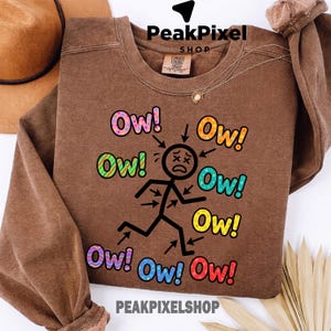Op de afbeelding: Bruin sweatshirt met de tekst "Ow!" in kleurrijke bubbelletters rond een stokfiguur met pijlen. De stokfiguur rent met een bedroefde uitdrukking. De merknaam "PeakPixel Shop" staat bovenaan en onderaan.