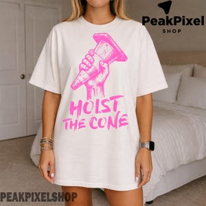 Pu&ograve; includere: T-shirt color crema con grafica rosa di una mano che tiene un cono stradale, con la scritta rosa "HOIST THE CONE" sotto. La maglietta ha una vestibilit&agrave; ampia.