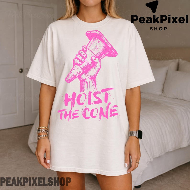 Pu&ograve; includere: T-shirt color crema con grafica rosa di una mano che tiene un cono stradale, con la scritta rosa "HOIST THE CONE" sotto. La maglietta ha una vestibilit&agrave; ampia.