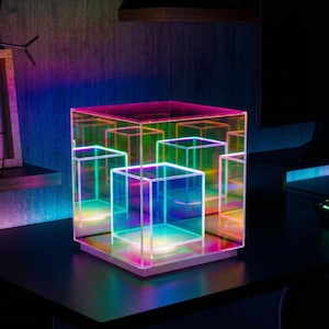 Puede incluir: Una taza de cerámica blanca con un asa negra y el texto "But First, Coffee" en negrita negra. La taza se encuentra sobre un fondo borroso, sugiriendo un ambiente acogedor.