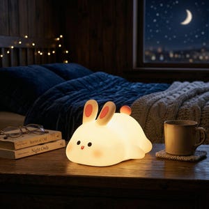 May include: A soft, glowing bunny-shaped night light with pink accents on its ears and cheeks sits on a wooden table. The lamp is next to a stack of books and a mug. The background includes a bed and a window with a night sky view.
