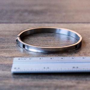Könnte beinhalten: Ein silberfarbenes Armreif-Armband mit rechteckigem Verschluss liegt auf einer Holzoberfläche neben einem Lineal. Das Armband hat ein schlichtes, kreisförmiges Design. Das Lineal ist weiß mit schwarzen Markierungen.