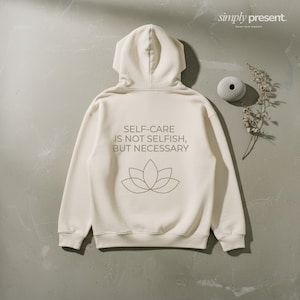 Könnte beinhalten: Cremefarbener Kapuzenpullover mit Kapuze und dem Text "SELF-CARE IS NOT SELFISH, BUT NECESSARY" sowie einer Lotusblumen-Grafik auf dem Rücken. Der Hoodie wird auf einer neutralen Oberfläche mit dekorativen Elementen präsentiert.