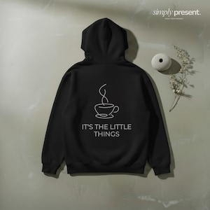Op de afbeelding: Zwarte hoodie met een witte koffiekopjesafbeelding en de tekst "IT'S THE LITTLE THINGS". De hoodie wordt getoond tegen een neutrale achtergrond, met een klein decoratief item en bladeren.