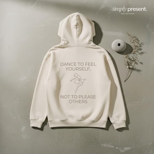 Könnte beinhalten: Cremefarbener Kapuzenpullover mit Kordelzug. Auf der Rückseite steht der Text "DANCE TO FEEL YOURSELF, NOT TO PLEASE OTHERS" mit einer Strichzeichnung einer Tänzerin. Der Hoodie wird auf einer strukturierten Oberfläche gezeigt.