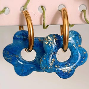 May include: A pair of gold hoop earrings with blue flower-shaped pendants. The pendants have a marbled effect with gold flecks. The earrings are hanging on a pink surface with green vines.