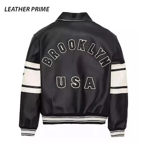 May include: A black varsity jacket with white stripes on the sleeves and ribbed cuffs. The back features the text BROOKLYN in an arched design above the text USA in white lettering with black outlines. The text LEATHER PRIME appears in the top corner.