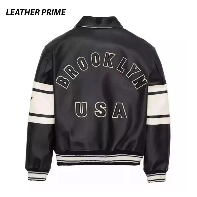 May include: A black varsity jacket with white stripes on the sleeves and ribbed cuffs. The back features the text BROOKLYN in an arched design above the text USA in white lettering with black outlines. The text LEATHER PRIME appears in the top corner.
