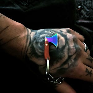 May include: A miniature axe keychain featuring a vibrant, iridescent rainbow-colored metal blade and a smooth, brown wooden handle. The small axe is attached to a silver-toned metal split ring and a short, sturdy link chain for secure attachment.