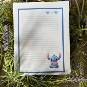 Pode incluir: Um bloco de notas pautado com uma borda azul e três ícones de coração na parte superior. Um personagem de desenho animado segurando um coração está na parte inferior. O bloco de notas é cercado por vegetação.