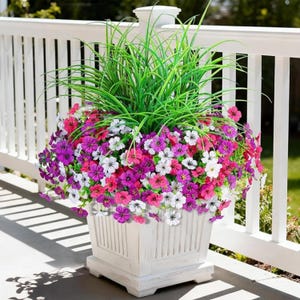 May include: A square, white wooden planter filled with vibrant flowers and tall green grass. The flowers are a mix of pink, purple, and white, creating a colorful display. The planter is on a porch with a white railing.