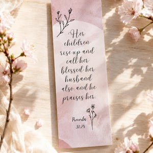Könnte beinhalten: Ein rechteckiger Lesezeichen mit einem rosa Aquarellhintergrund und schwarzer Schrift. Der Text lautet: "Ihre Kinder stehen auf und preisen sie glücklich, auch ihr Mann, und er lobt sie. Sprüche 31:28." Florale Illustrationen oben und unten.