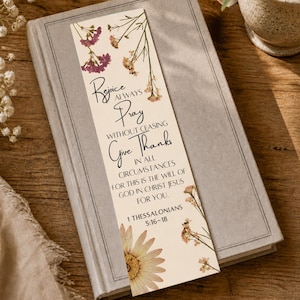 Könnte beinhalten: Ein beiger Lesezeichen mit floralen Akzenten und dem Text "Freut euch allezeit, betet ohne Unterlass, seid dankbar in allen Dingen". Das Lesezeichen liegt auf einem geschlossenen Buch mit grauem Einband. Der Text enthält den Bibelvers 1. Thessalonicher 5:16-18.
