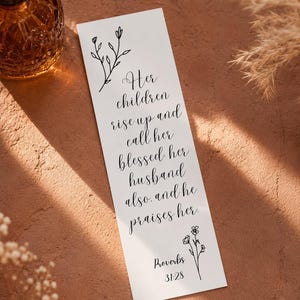 Könnte beinhalten: Ein weißes Lesezeichen mit schwarzer Schrift: „Ihre Kinder stehen auf und preisen sie glücklich, auch ihr Mann, und er lobt sie. Sprüche 31:28.“ Mit floralen Illustrationen.
