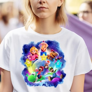 Może przedstawiać: Biała koszulka z kolorową grafiką postaci z Super Mario. Projekt przedstawia Mario, Luigiego, Księżniczkę Peach, Toada i Yoshiego na kosmicznym tle z gwiazdami.