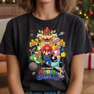 Może przedstawiać: Czarny t-shirt z kolorową grafiką postaci z gry Super Mario Galaxy. Projekt zawiera Mario, Bowsera, Księżniczkę Peach, Yoshiego i inne postacie, z logo gry na dole.