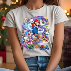 Może przedstawiać: Jasnoszara koszulka z kolorową grafiką z filmu "The Super Mario Galaxy Movie". Projekt przedstawia Mario, Księżniczkę Rosalina i r&oacute;żne postacie z gry, otoczone wirującą, fioletowo-niebieską galaktyką.