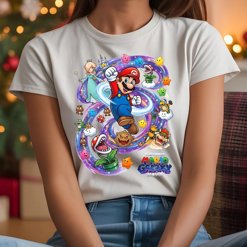 Może przedstawiać: Jasnoszara koszulka z kolorową grafiką z filmu "The Super Mario Galaxy Movie". Projekt przedstawia Mario, Księżniczkę Rosalina i r&oacute;żne postacie z gry, otoczone wirującą, fioletowo-niebieską galaktyką.