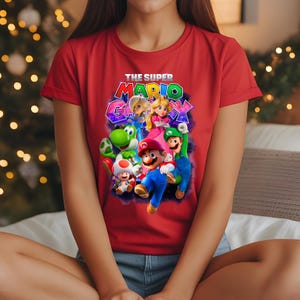 Może przedstawiać: Czerwona koszulka z grafiką postaci z gry wideo Super Mario Galaxy. Projekt zawiera Mario, Luigiego, księżniczkę Peach i inne postacie. Tekst "THE SUPER MARIO GALAXY" jest wydrukowany nad postaciami.