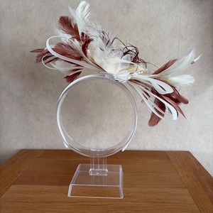 May include: A decorative headpiece featuring a cluster of cream and dark brown feathers arranged on a thin, clear plastic headband. The design includes wispy plumes, looped ribbon accents, and small white beads, all mounted on a clear stand.