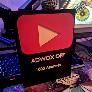 May include: A black and red YouTube play button award with the text "ADWOX OFF" and "1000 Abonnés". A silver microphone rests on top of the award. The award is on a desk with a keyboard and computer screen in the background.