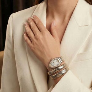 May include: A silver wristwatch with a white face and diamond detailing, worn with a matching silver bracelet. The person is wearing a cream-coloured blazer. The watch and bracelet are wrapped around the wrist.