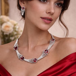 Könnte beinhalten: Elegante Halskette und passende Ohrringe aus Silber mit großen, ovalen, roten Edelsteinen. Der Schmuck ist mit kleinen, funkelnden Steinen besetzt. Die Frau trägt ein rotes Kleid.
