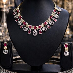 May include: A gold necklace and earring set with ruby-red and clear crystal stones, displayed on a black bust. The necklace features oval and teardrop-shaped red stones, with clear crystals. The earrings mirror the necklace design.