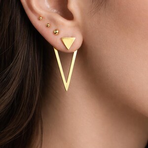 Puede incluir: Primer plano de pendientes dorados. Los pendientes incluyen tres pequeños tachuelas de bola y un par de pendientes geométricos con una tachuela triangular y una gran forma de V invertida. Las joyas se llevan en una oreja.