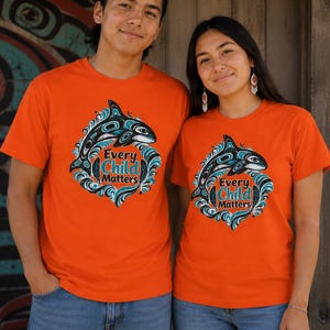 May include: Two people wearing orange t-shirts featuring a black and teal orca whale design. The text "Every Child Matters" is below the whale. The shirts are paired with blue jeans.