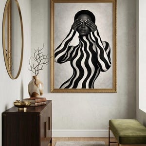May include: Black and white portrait of a person with their eyes covered, wearing a high-necked top and body paint with a bold, wavy stripe pattern. The artwork is framed in gold and hangs in a room with a gold mirror and a dark wood cabinet.