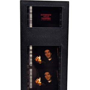 May include: A vertical black display frame containing a film strip from Interview with the Vampire. The strip features five sequential frames of a person holding a lit candle, topped with a header displaying the movie title in red text.