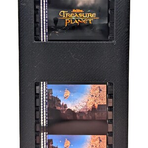 May include: A black film strip holder with two film strips. The top film strip displays the title "Treasure Planet" in gold lettering. The bottom film strips show a scene with a ship sailing over a city.