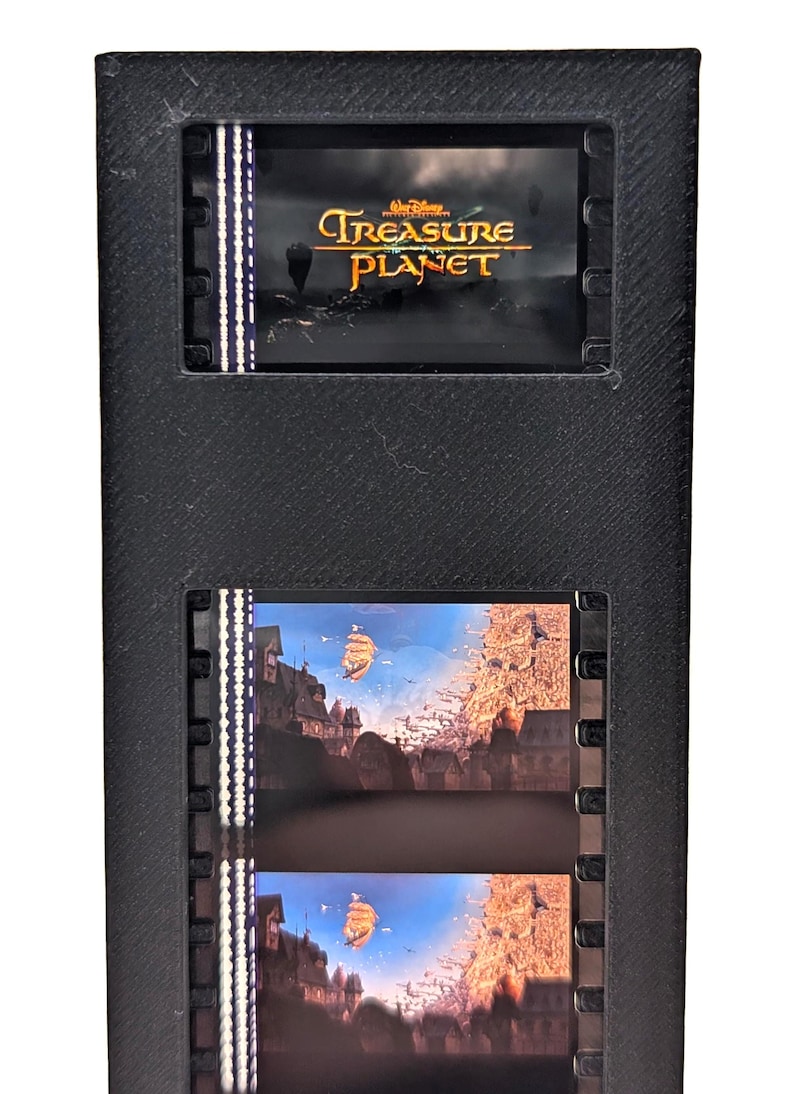 May include: A black film strip holder with two film strips. The top film strip displays the title "Treasure Planet" in gold lettering. The bottom film strips show a scene with a ship sailing over a city.