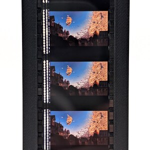 May include: A black film strip holder containing five frames of a fantasy landscape. Each frame shows a ship sailing towards a town with tall buildings under a blue sky. The scene is illuminated by warm, golden light.