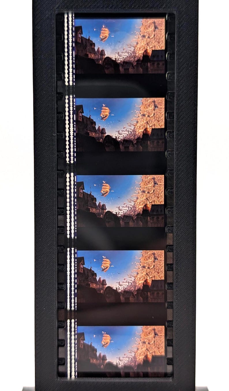 May include: A black film strip holder containing five frames of a fantasy landscape. Each frame shows a ship sailing towards a town with tall buildings under a blue sky. The scene is illuminated by warm, golden light.