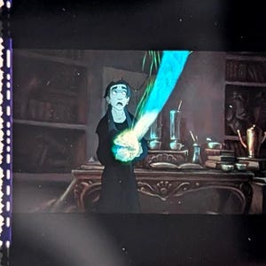 Puede incluir: Celda de pel&iacute;cula con una escena de una pel&iacute;cula animada. Un personaje vestido de negro sostiene un objeto azul brillante. El fondo muestra una habitaci&oacute;n con un escritorio, libros y varios objetos.