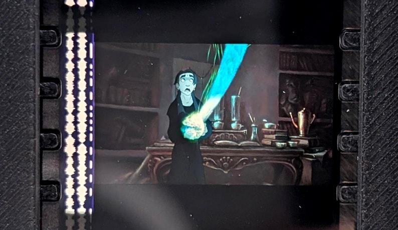 Puede incluir: Celda de pel&iacute;cula con una escena de una pel&iacute;cula animada. Un personaje vestido de negro sostiene un objeto azul brillante. El fondo muestra una habitaci&oacute;n con un escritorio, libros y varios objetos.