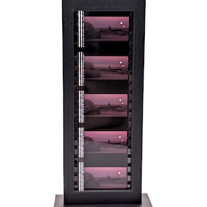 May include: A vertical black rectangular display frame containing a strip of five identical 35mm film frames. Each frame shows a silhouette of a person standing in a desert landscape under a pink sky with two white circular light spots.