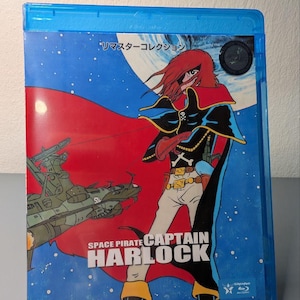 Peut inclure: Un boîtier Blu-ray bleu avec l'anime Space Pirate Captain Harlock. L'illustration de la couverture représente le personnage Harlock avec une cape rouge, un vaisseau spatial et le titre. Le boîtier repose sur un objet rectangulaire rouge.