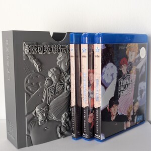 Op de afbeelding: Een grijze boxset met reliëfkunst en Japanse tekst, met verschillende Blu-ray-cases. De cases hebben blauwe ruggen en bevatten illustraties en tekst in het Japans en Engels, waaronder "Legend of the Galactic Heroes."