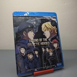 Puede incluir: Una caja Blu-ray azul con personajes de anime de "Legend of the Galactic Heroes". La portada muestra a dos hombres enfrentados, con el título en inglés y japonés. La caja está sujeta por un soporte rojo.