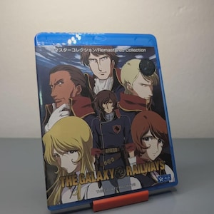 Può includere: Una custodia Blu-ray blu con personaggi anime e il titolo "THE GALAXY RAILWAYS". La copertina presenta diversi personaggi in uniformi militari. La custodia è supportata da un piccolo supporto rosso.
