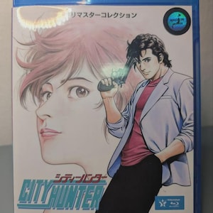 Peut inclure: Un boîtier Blu-ray bleu avec une illustration de style manga. La couverture présente un homme tenant une arme à feu, avec le visage d'une femme en arrière-plan. Le titre "CITY HUNTER" est affiché en bleu et vert. Contient du texte japonais.