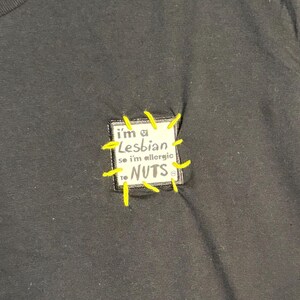 May include: Black t-shirt with a sewn-on patch. The patch reads "I'm a Lesbian so I'm allergic to NUTS" in a handwritten style. The patch is stitched with yellow thread.