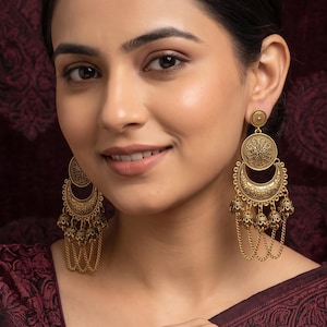May include: A pair of gold-toned dangle earrings featuring a circular stud connected to a crescent-shaped piece with intricate embossed patterns and dangling chain elements that terminate in small, bell-shaped charms with a matte finish.