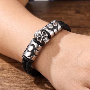 Op de afbeelding: Een armband van gevlochten zwart leer met drie zilverkleurige metalen kralen. De middelste kraal heeft de vorm van een schedel, terwijl de twee zijdelingse kralen een glad, gebogen en abstract metalen ontwerp hebben rond het leren koord.