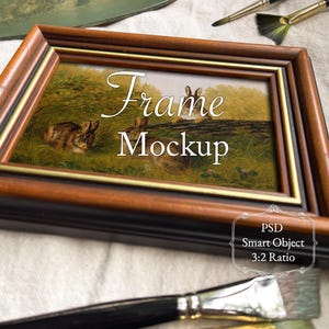 May include: A framed artwork featuring a painting of rabbits in a field, with the text "Frame Mockup". The frame is brown with a gold inner border. Paintbrushes and a palette are in the background. The image also includes the text "PSD Smart Object 3:2 Ratio".